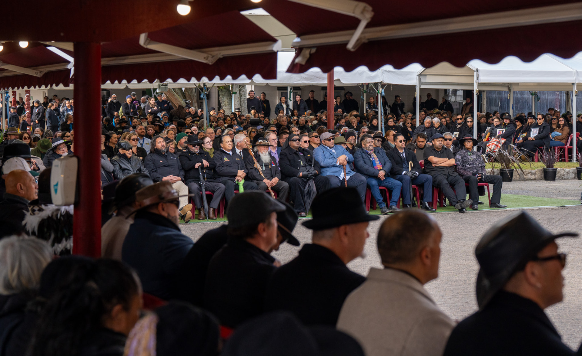 A large crowd gathered together at a community hui under red marquees