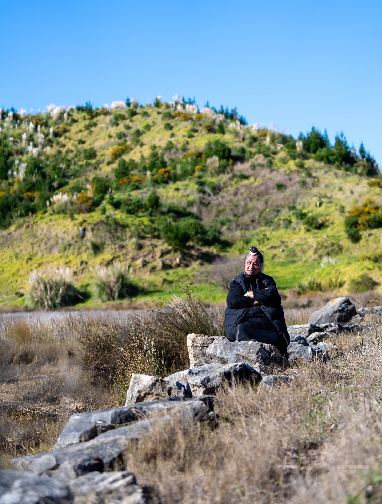 A kuia sitting on a rocky outcrop, surrounded by native bush and rolling hills