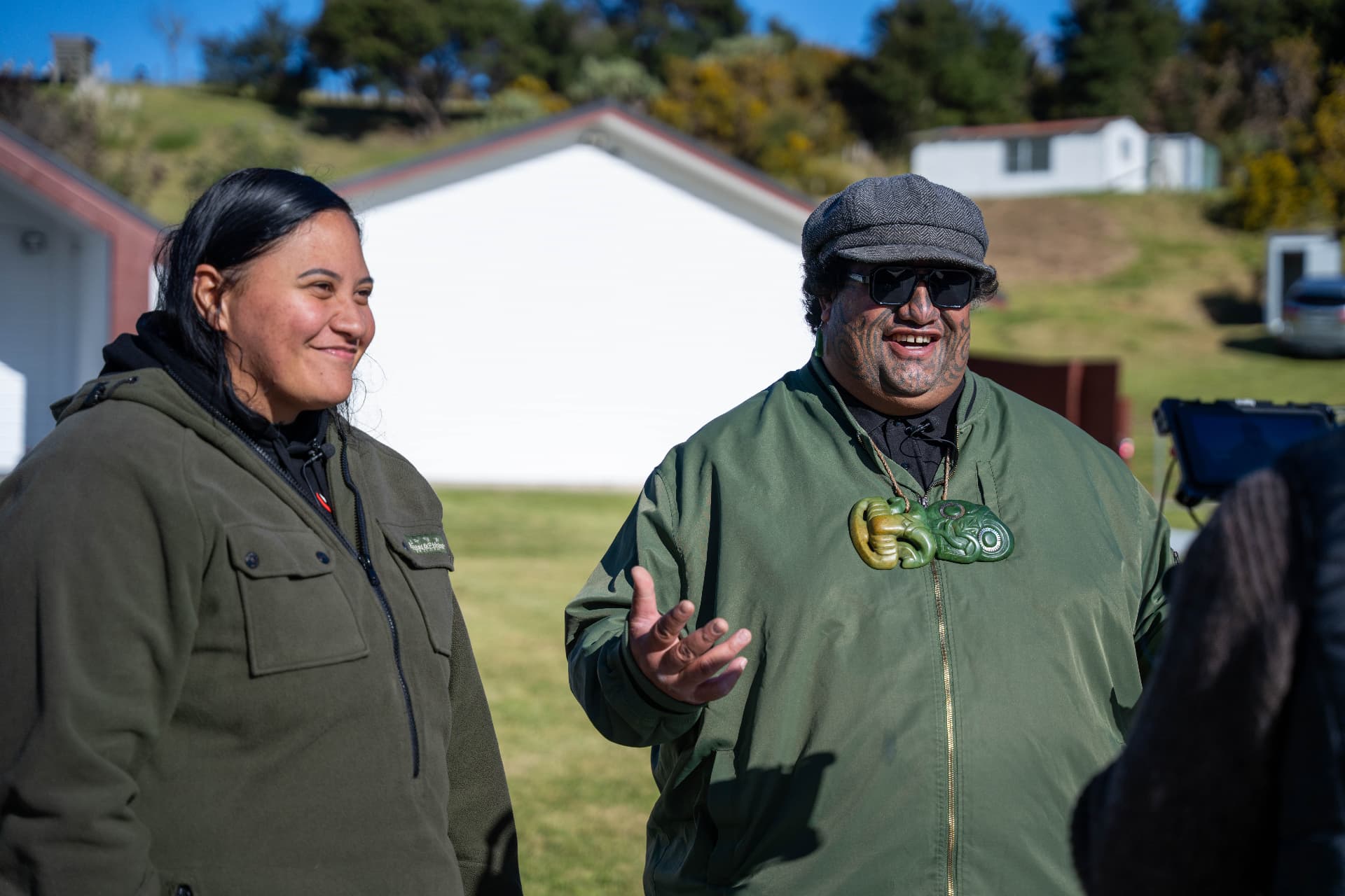 Two people sharing stories outdoors, one wearing tā moko and a pounamu pendant